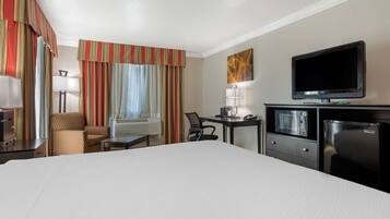 A hotel room with a large bed, a desk with a lamp, a television, a refrigerator, and a chair.