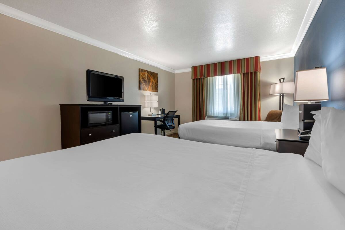 A hotel room with two beds, a desk with a TV, a lamp, and a window with curtains.