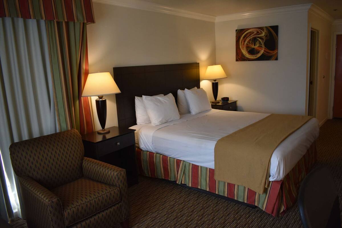 A hotel room with a bed, two bedside lamps, a chair, a nightstand, and a wall painting.