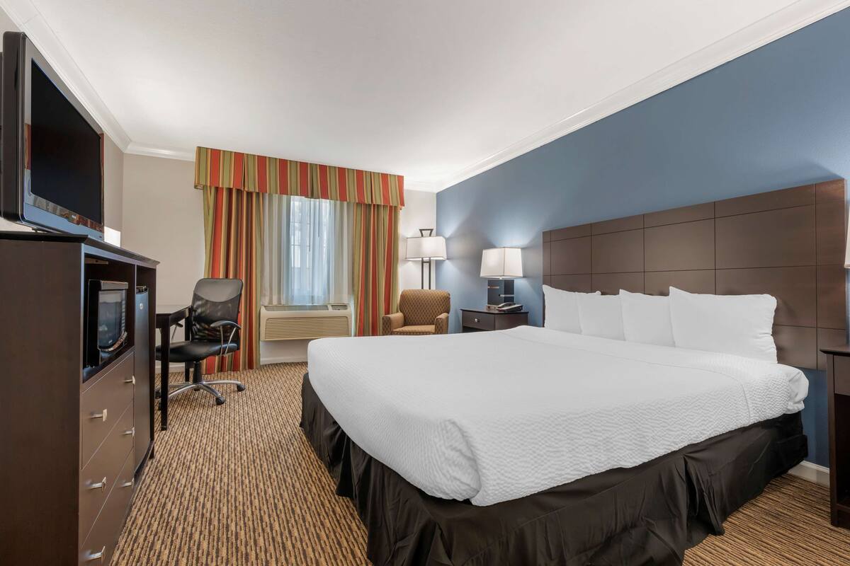 A hotel room with a large bed, a desk with a television, two chairs, a nightstand with lamps, and a window with curtains.