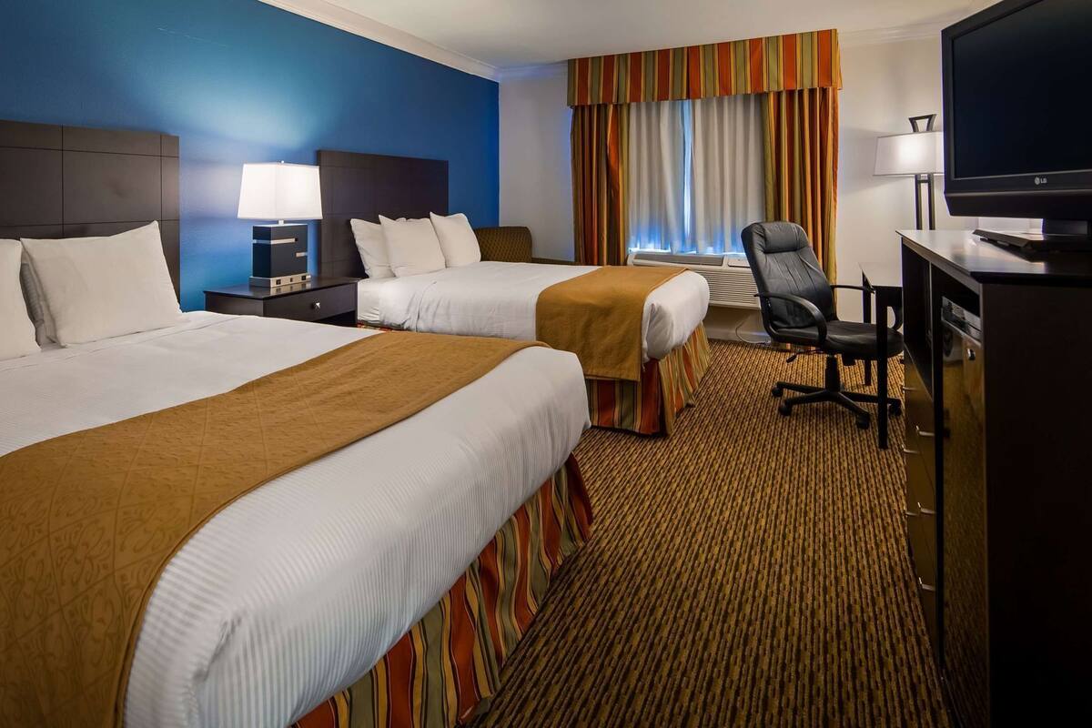 A hotel room with two beds, a desk with a chair, a TV, and a window with curtains.