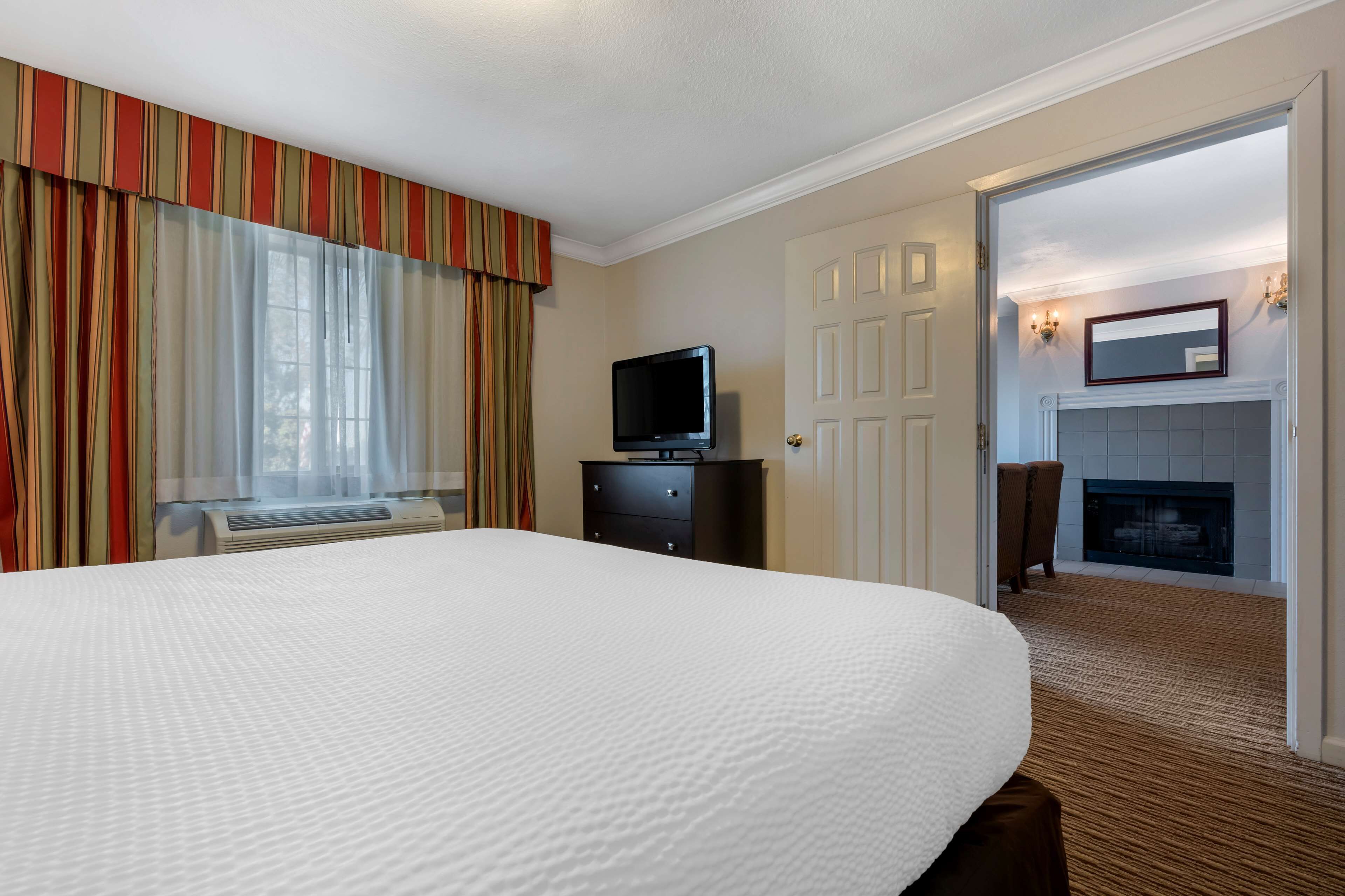 A hotel room with a large bed, a television on a dresser, a fireplace, and a view of the outside through a window with curtains.