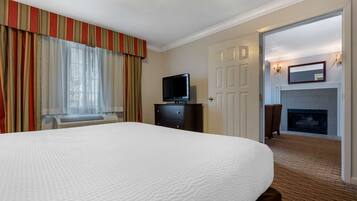 A hotel room with a large bed, a television on a dresser, a fireplace, and a view of the outside through a window with curtains.