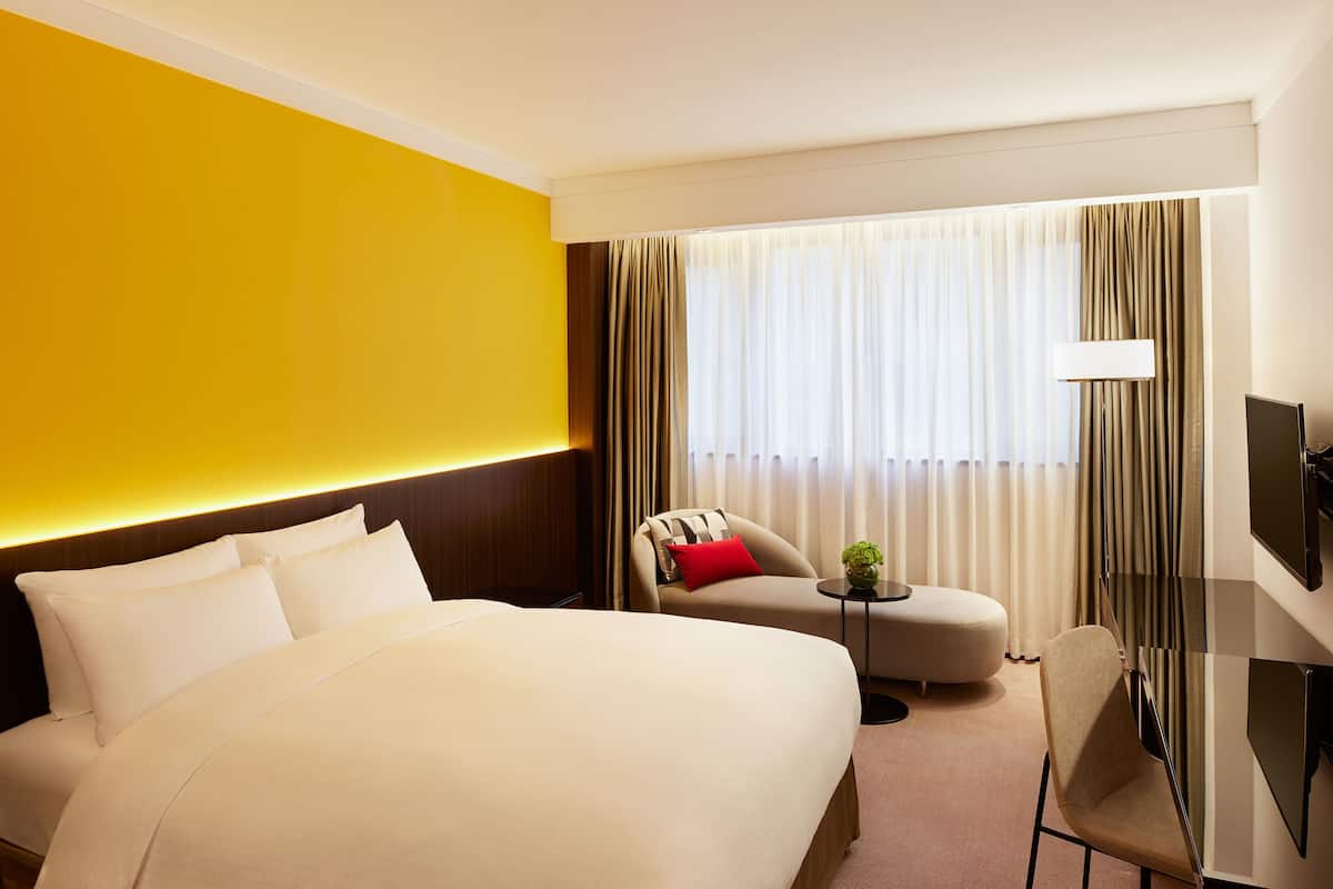 A hotel room with a large bed, a sofa, a small table with a plant, a TV, and a window with curtains.