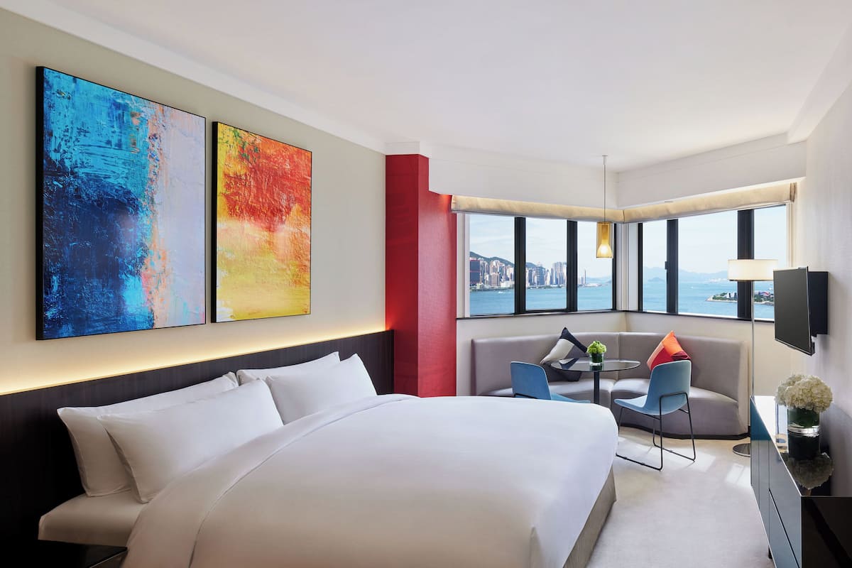 A modern hotel room with a large bed, a seating area with a table, and a city view through large windows.