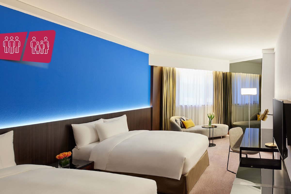 A hotel room with two beds, a desk, a TV, and a seating area with a view of the city.