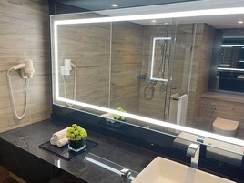 A modern bathroom with a large mirror, a glass-enclosed shower, a hairdryer, and a small plant on the counter.
