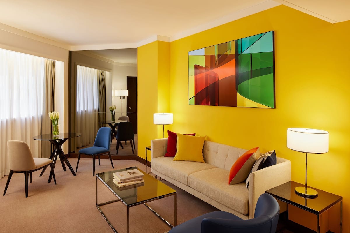 A modern hotel room with a yellow accent wall, a large abstract painting, a sofa with multiple cushions, a coffee table, a side table with a lamp, and a glass-top table with a vase of flowers.