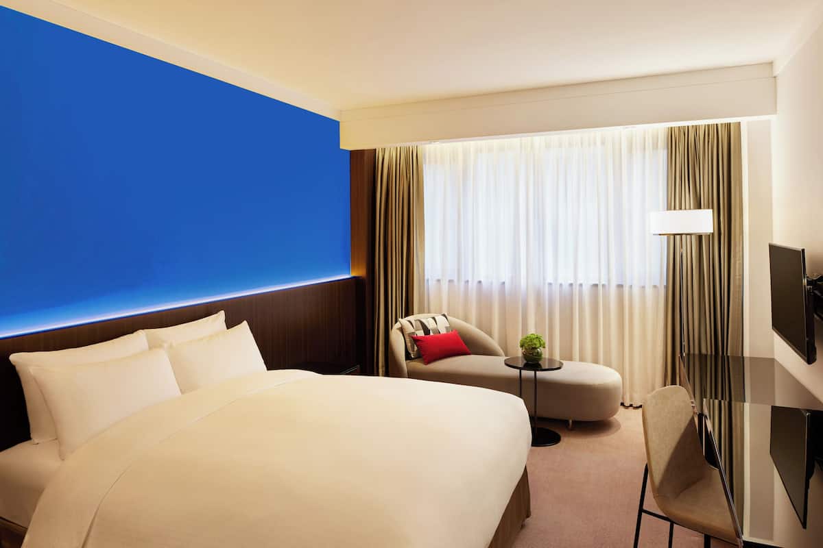 A hotel room with a large bed, a sofa, a small table with a plant, a TV, and a window with curtains.