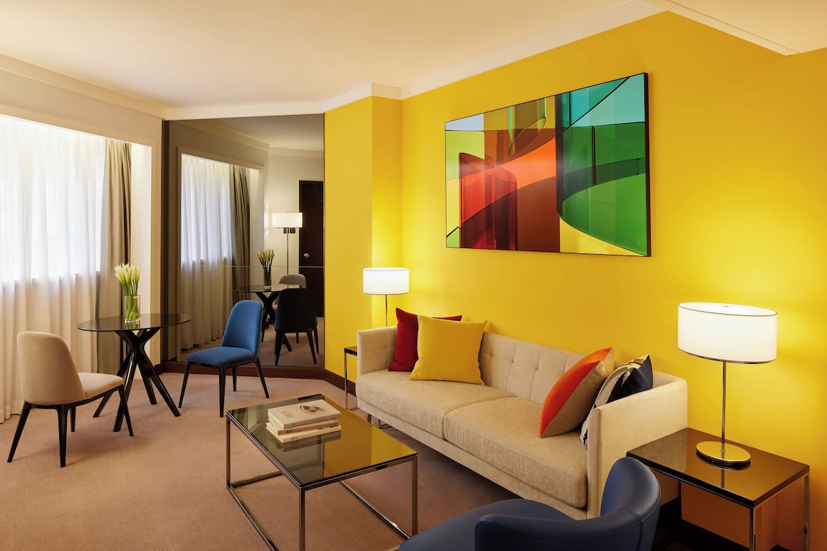 A modern hotel room with a yellow accent wall, a sofa, two armchairs, a coffee table, and a side table with a lamp.