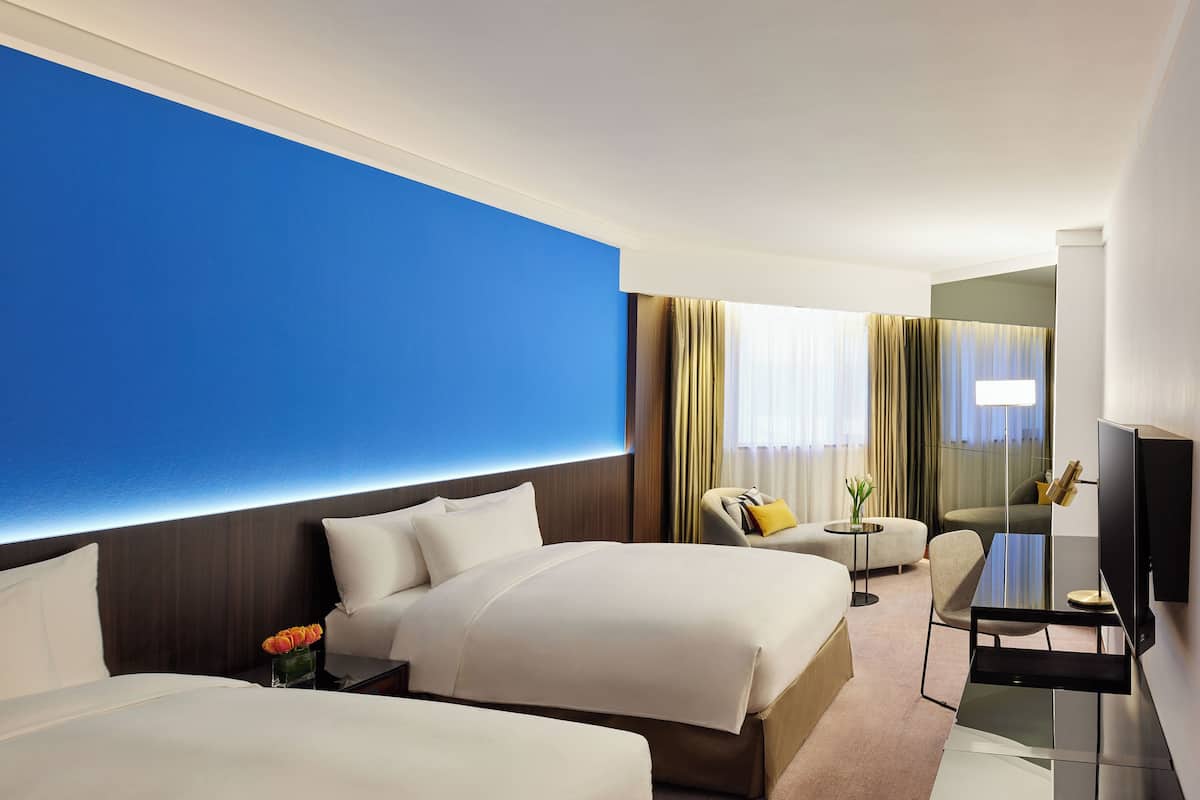 A hotel room with two beds, a desk, a chair, a TV, and a large blue accent wall.