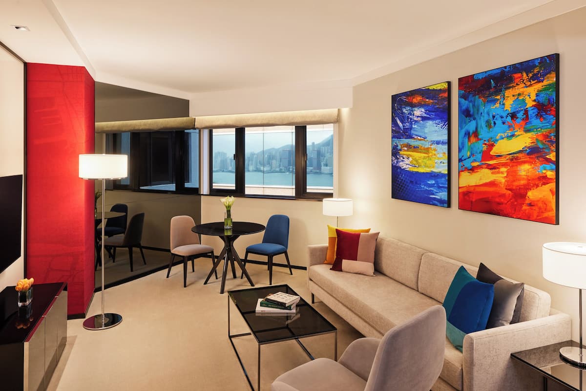 A modern living room with a sofa, coffee table, and chairs. There are two large windows with a city view.
