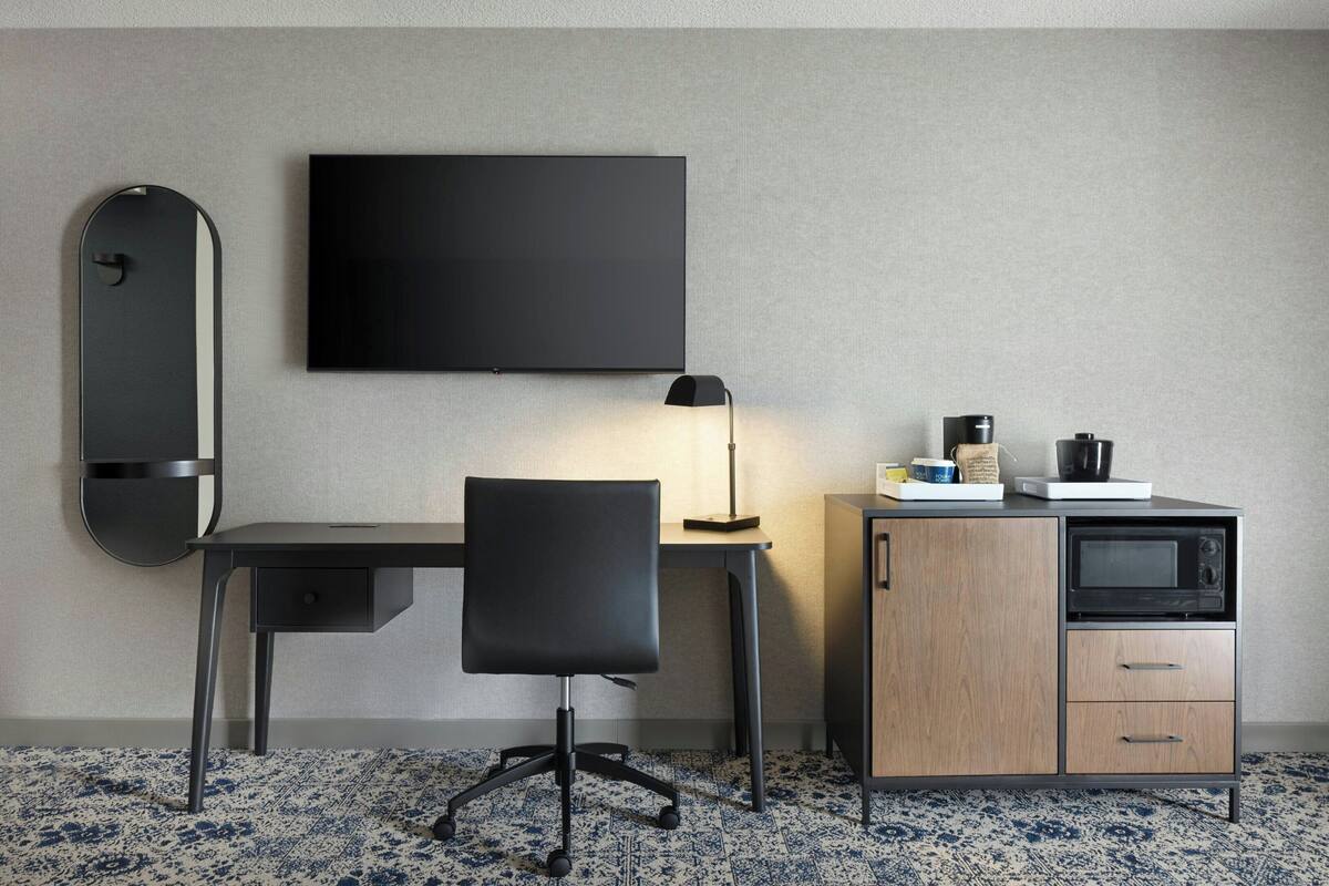 A modern hotel room with a desk, chair, flat-screen TV, lamp, microwave, and a cabinet.