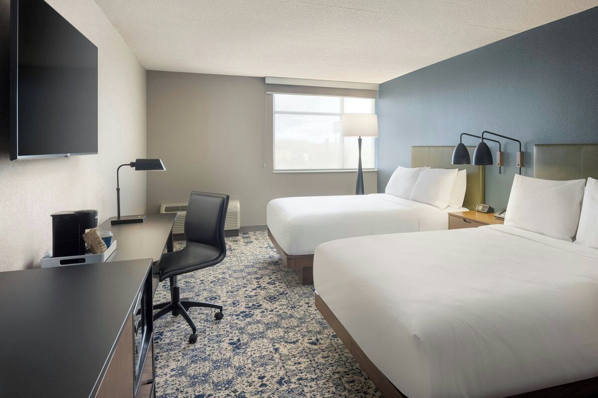 A hotel room with two beds, a desk, a chair, a TV, a lamp, and a window.