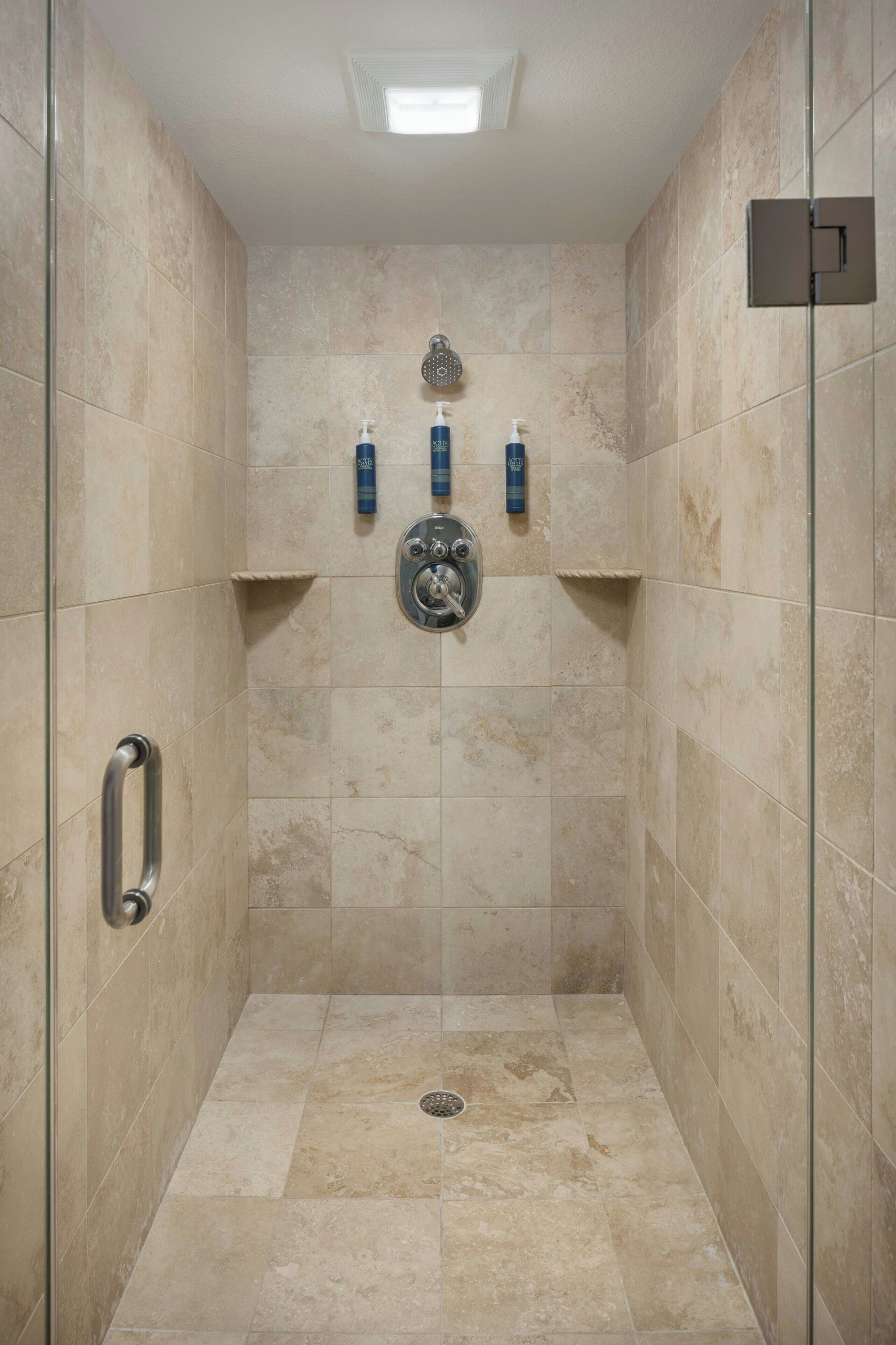 Eine bodenebene Dusche mit beigen Fliesen, einem Handbrausekopf und zwei Flaschen Shampoo und Conditioner an der Wand.