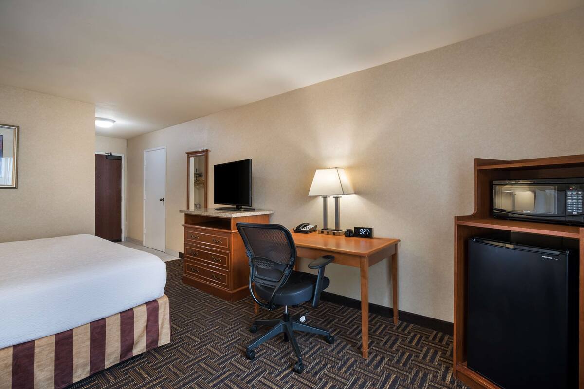 A hotel room with a bed, desk, chair, lamp, television, and refrigerator.