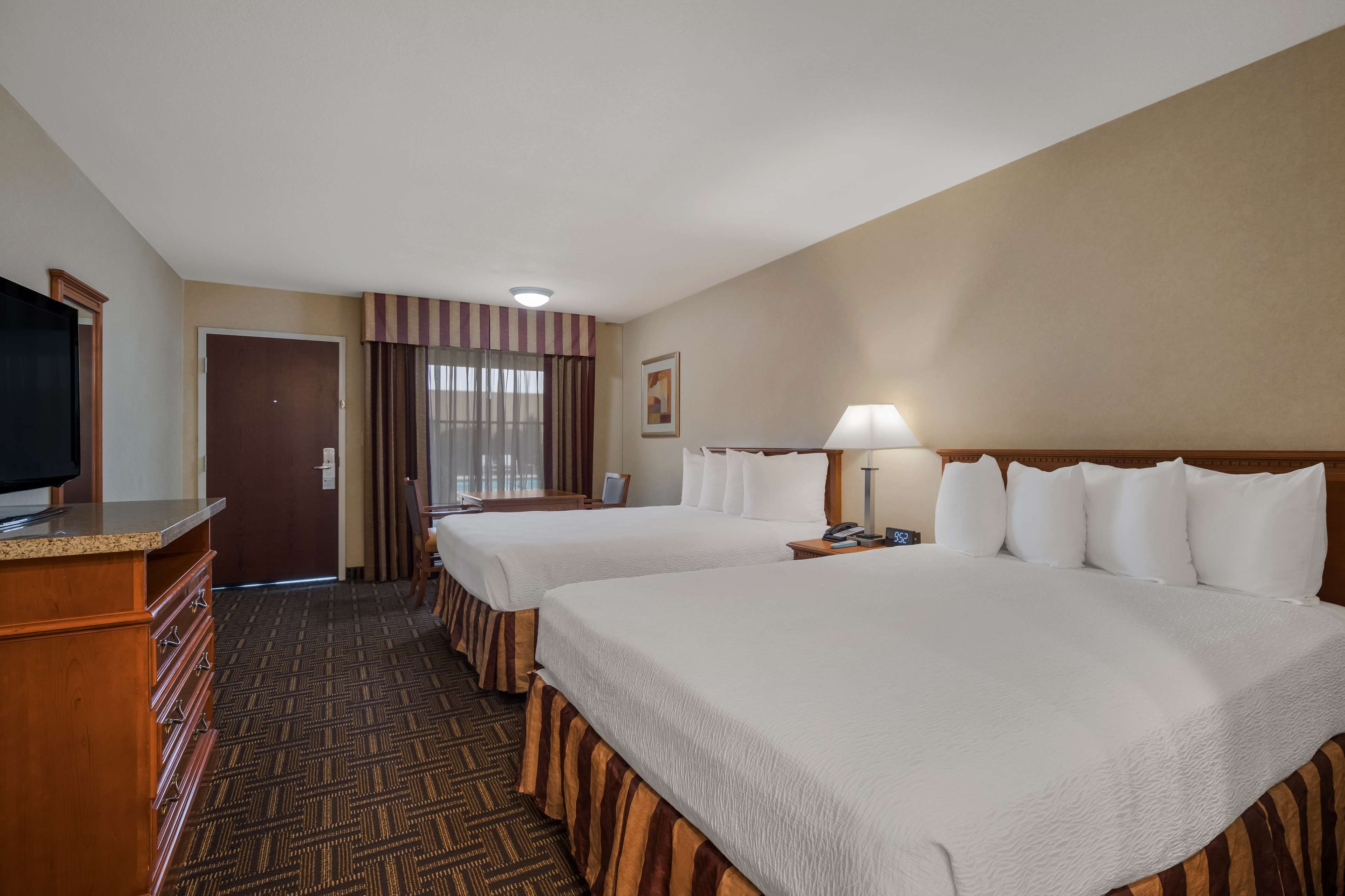 A hotel room with two beds, a television, a desk with drawers, a lamp, and a door with curtains.
