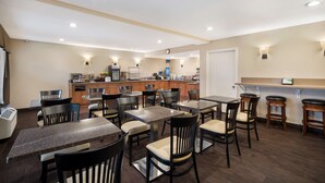 Een restaurantgedeelte van een hotel met meerdere tafels en stoelen, een keuken op de achtergrond en een bar met krukjes.