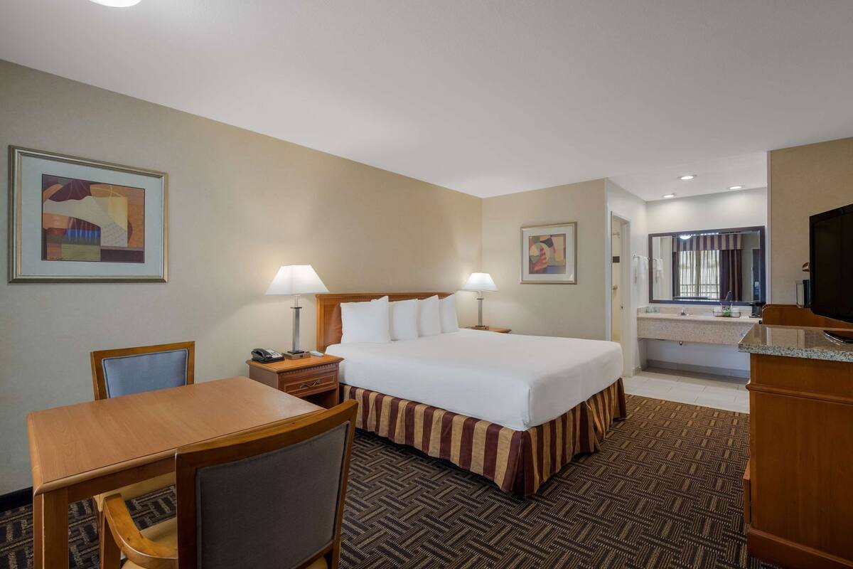 A hotel room with a bed, bedside tables, a desk, a chair, a TV, and a bathroom with a mirror and sink.