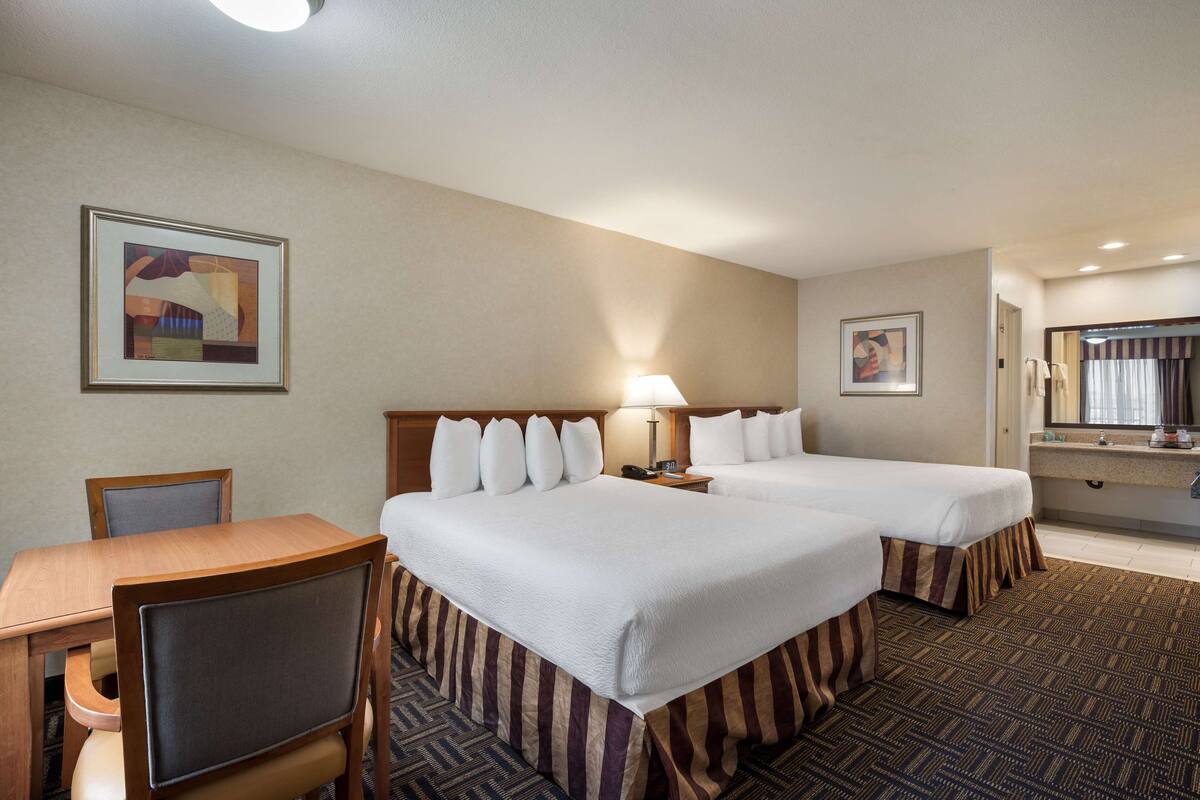 A hotel room with two beds, a desk, a chair, a lamp, and a bathroom with a mirror.