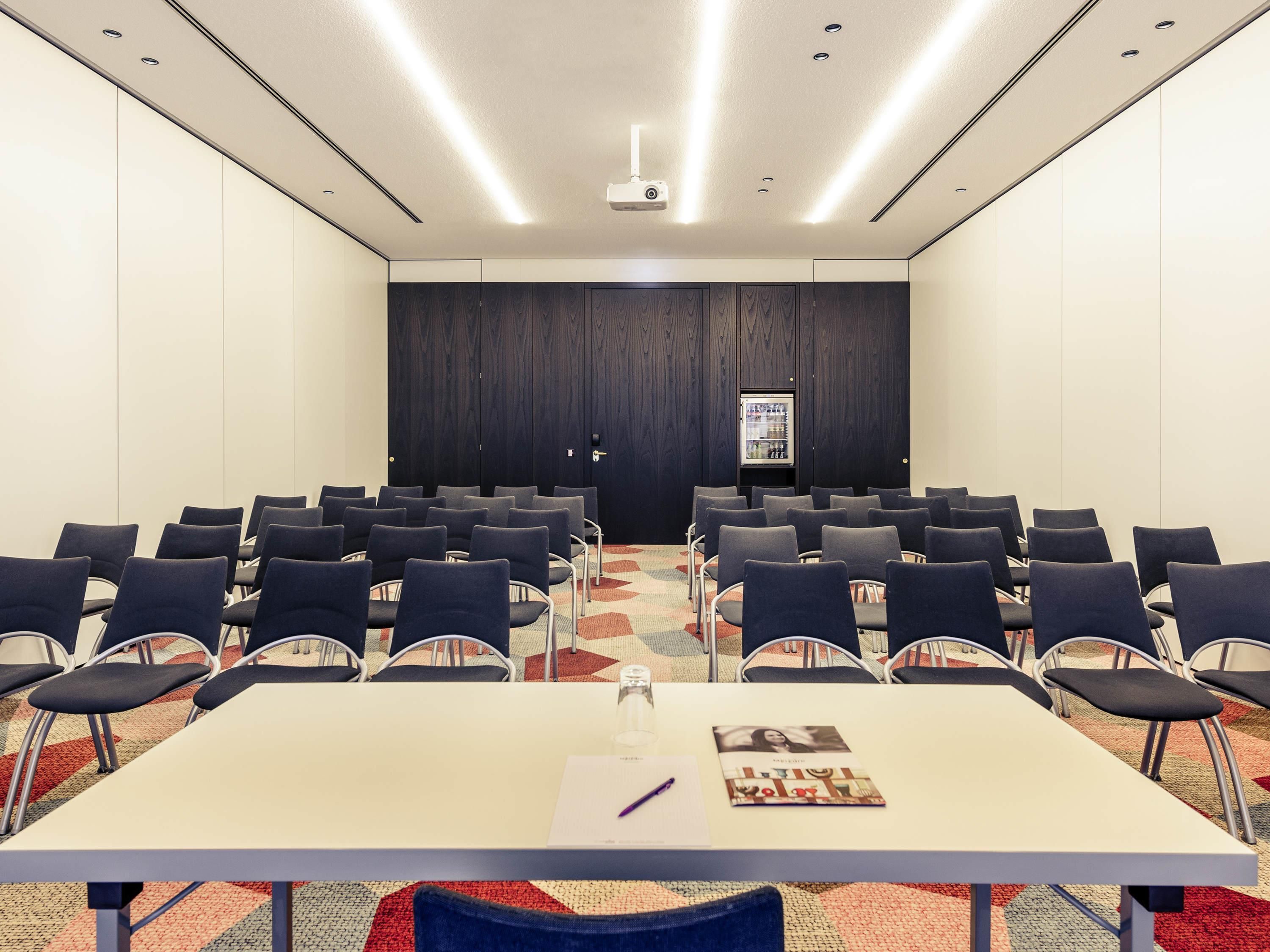 Une salle de conférence avec des rangées de chaises, une table avec un carnet et un stylo, un projecteur au plafond et un réfrigérateur rempli de boissons.