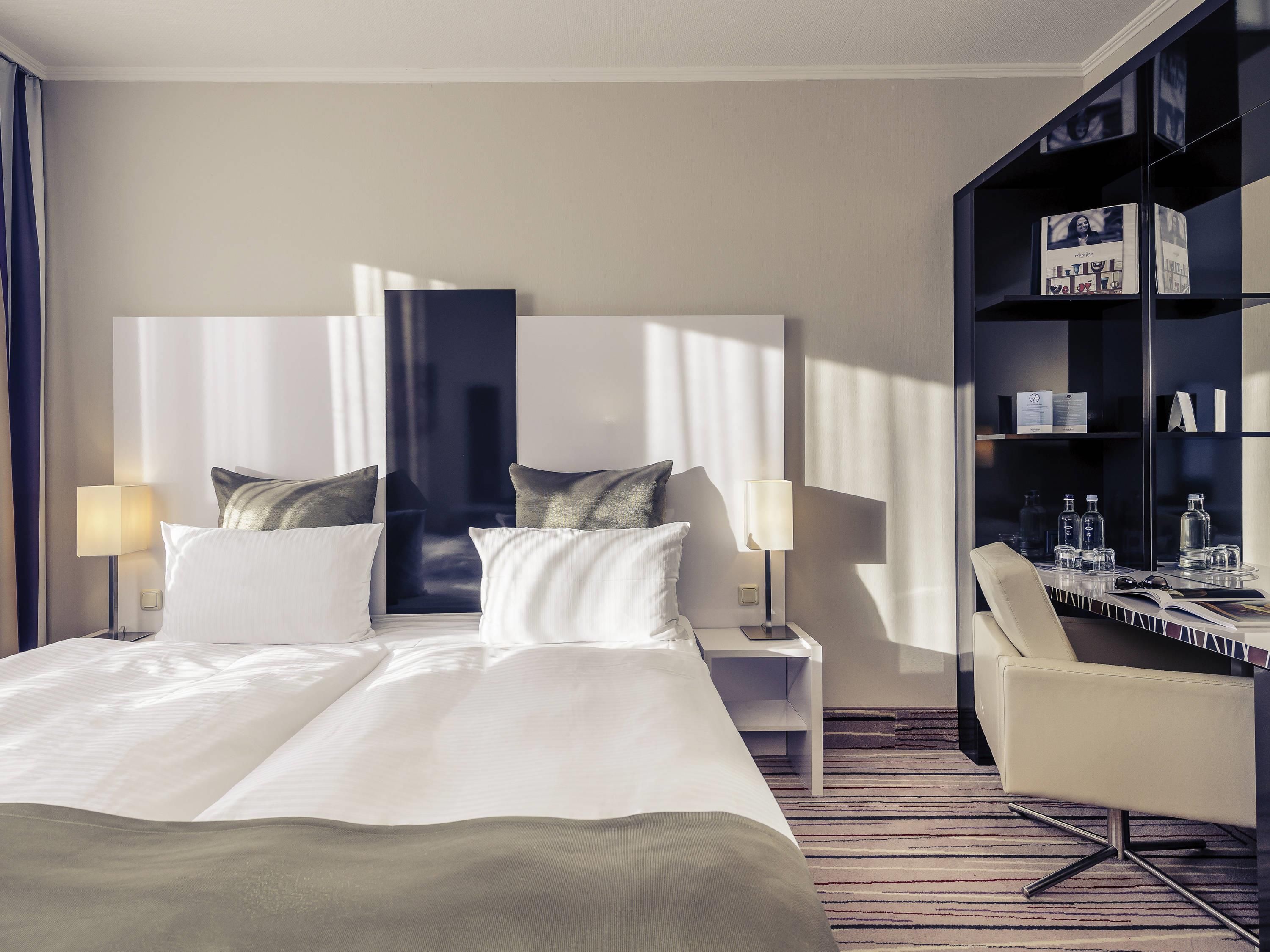 Une chambre d’hôtel avec un grand lit, deux tables de chevet, une chaise, une bibliothèque et une fenêtre avec des rideaux.