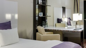 A hotel room with a bed, a desk with a chair, a minibar, a mirror, and a window with curtains. - Mercure Hotel Wiesbaden City (Wiesbaden)