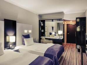 A hotel room with two beds, a desk, a chair, a mirror, and a door. - Mercure Hotel Wiesbaden City (Wiesbaden)