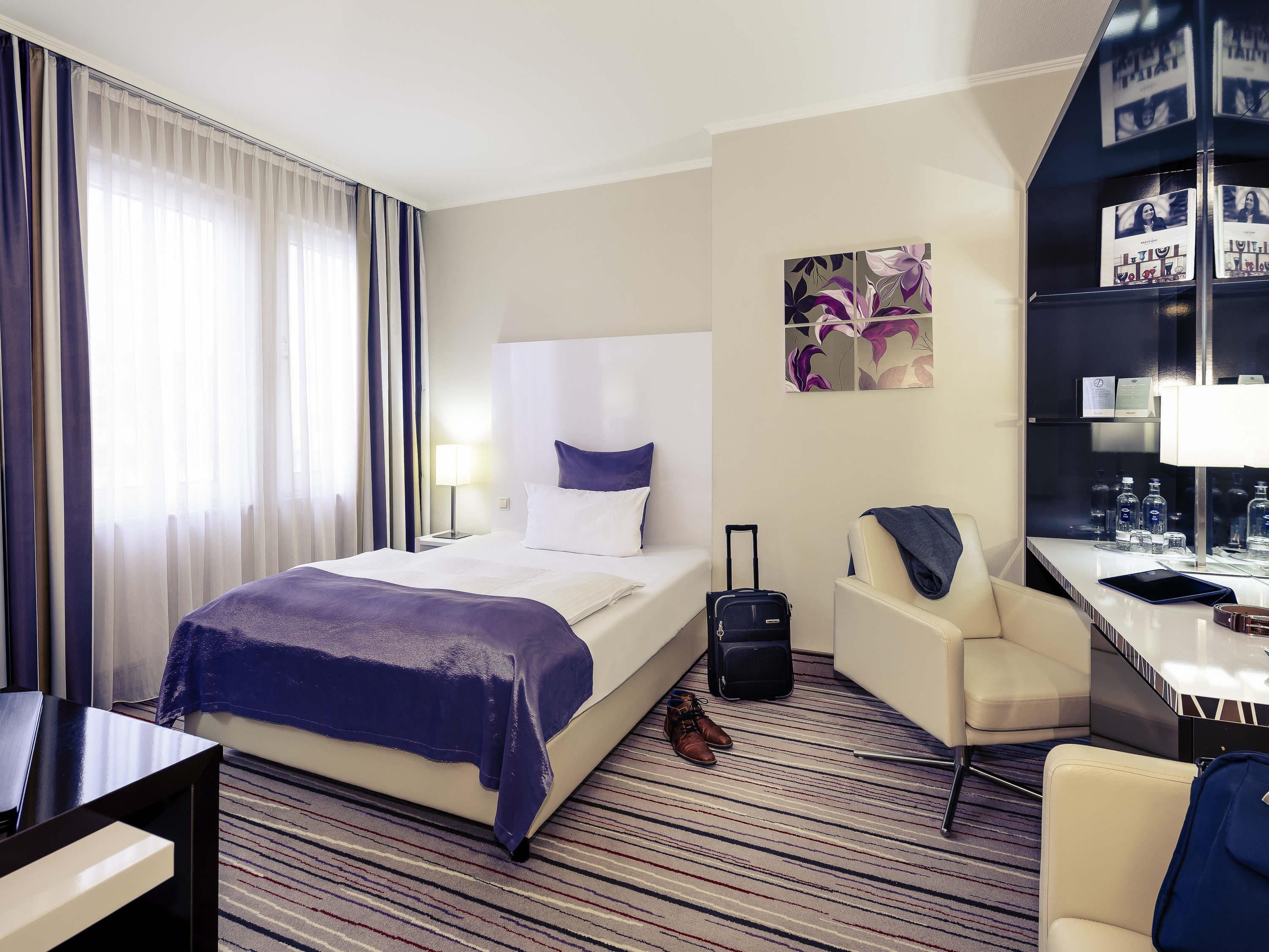 Une chambre d’hôtel avec un lit, un bureau, une chaise, une valise et une bibliothèque.