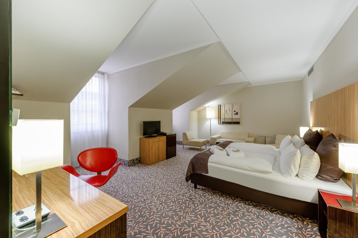 A hotel room with a bed, a desk, a red chair, a television, and a small table.