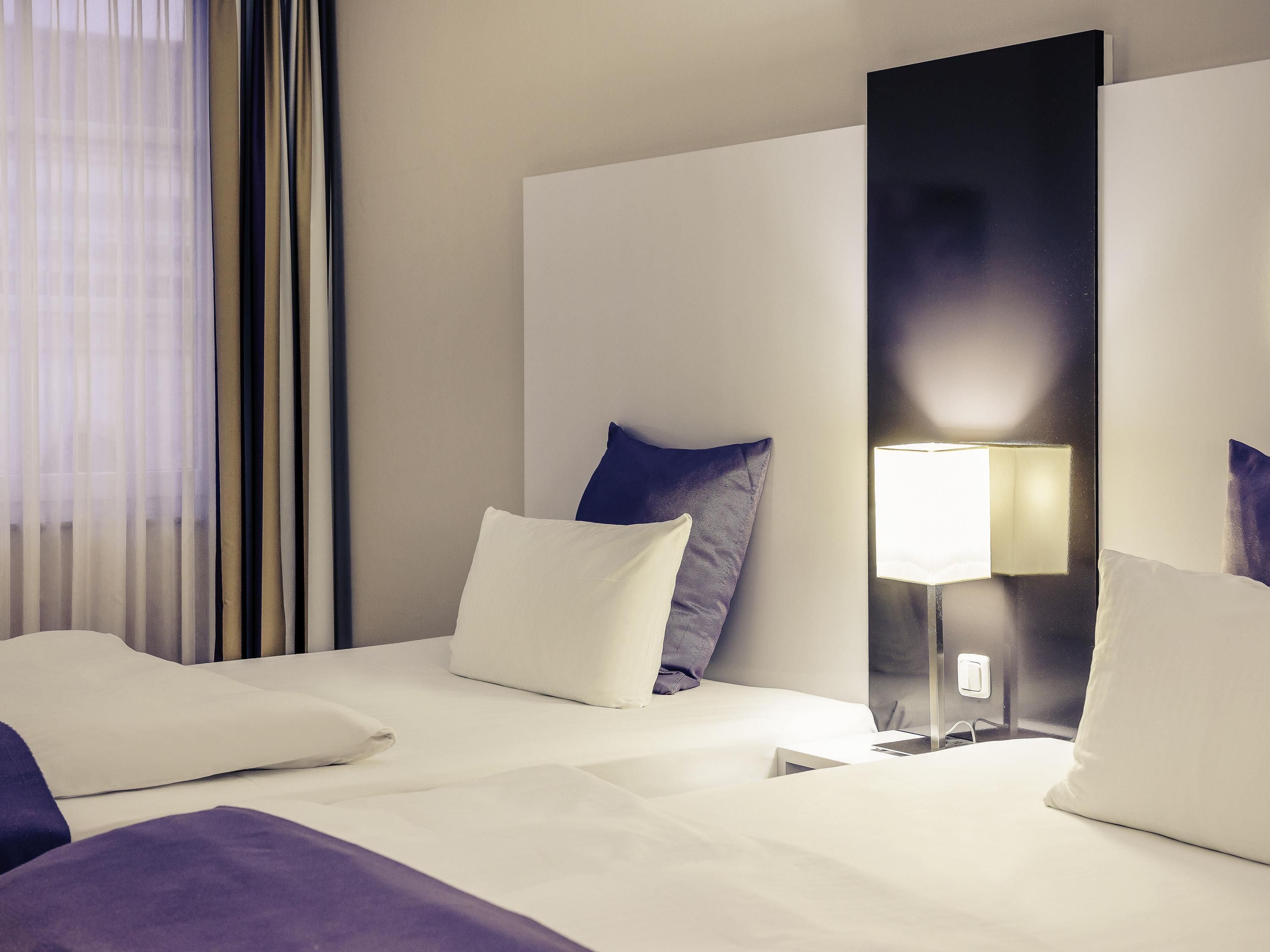Une chambre d’hôtel avec un lit, des oreillers, une table de chevet, une lampe et une fenêtre avec des rideaux.