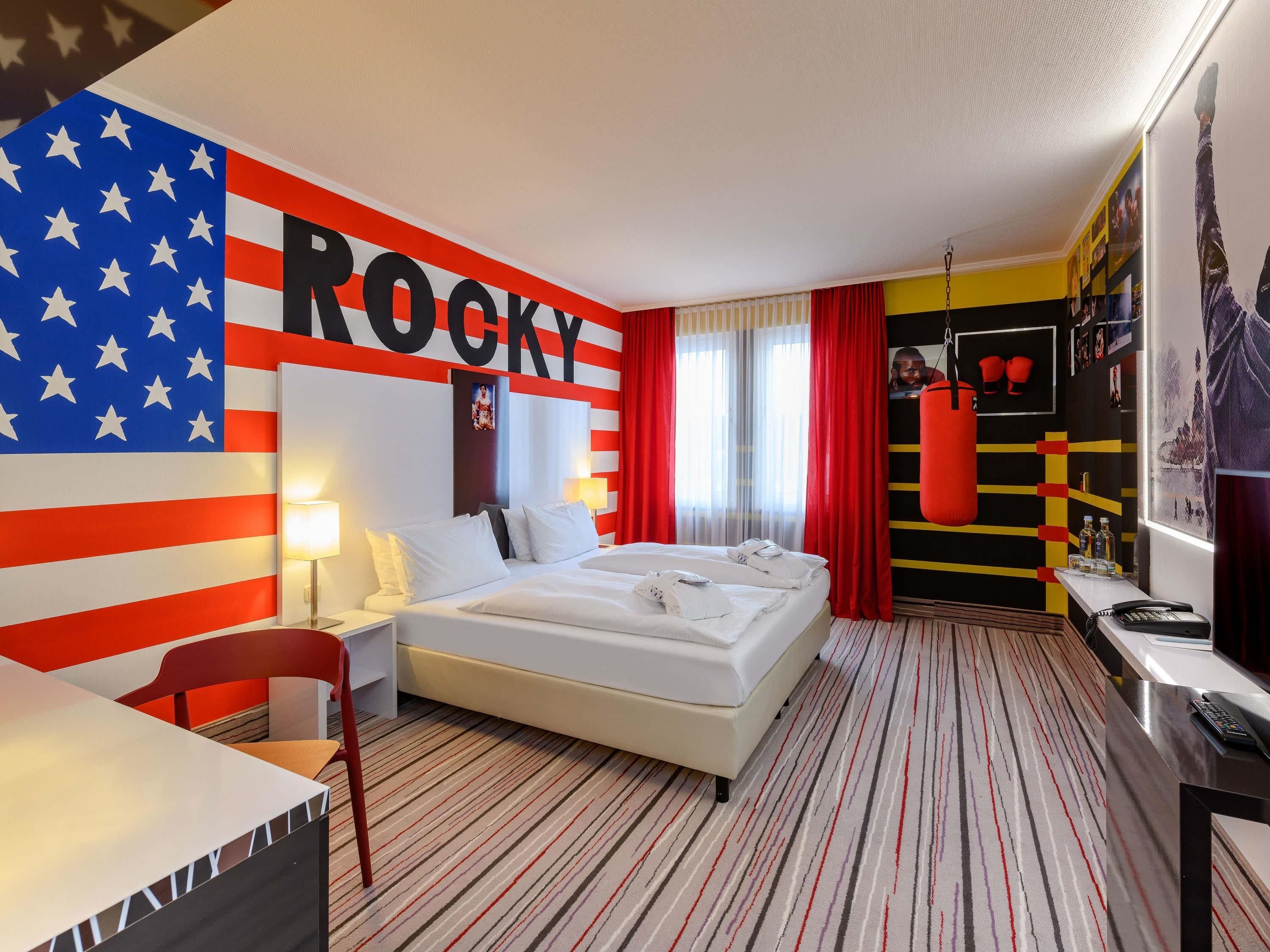 Ein Hotelzimmer mit einem Bett, einem Schreibtisch, einem Stuhl, einem Boxsack und einer Wand mit einem großen 'ROCKY'-Schild.