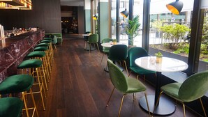 A bar area with green bar stools and a marble counter. There are round tables with green chairs and a large window with a view of the outside. - Mercure Hotel Wiesbaden City (Wiesbaden)