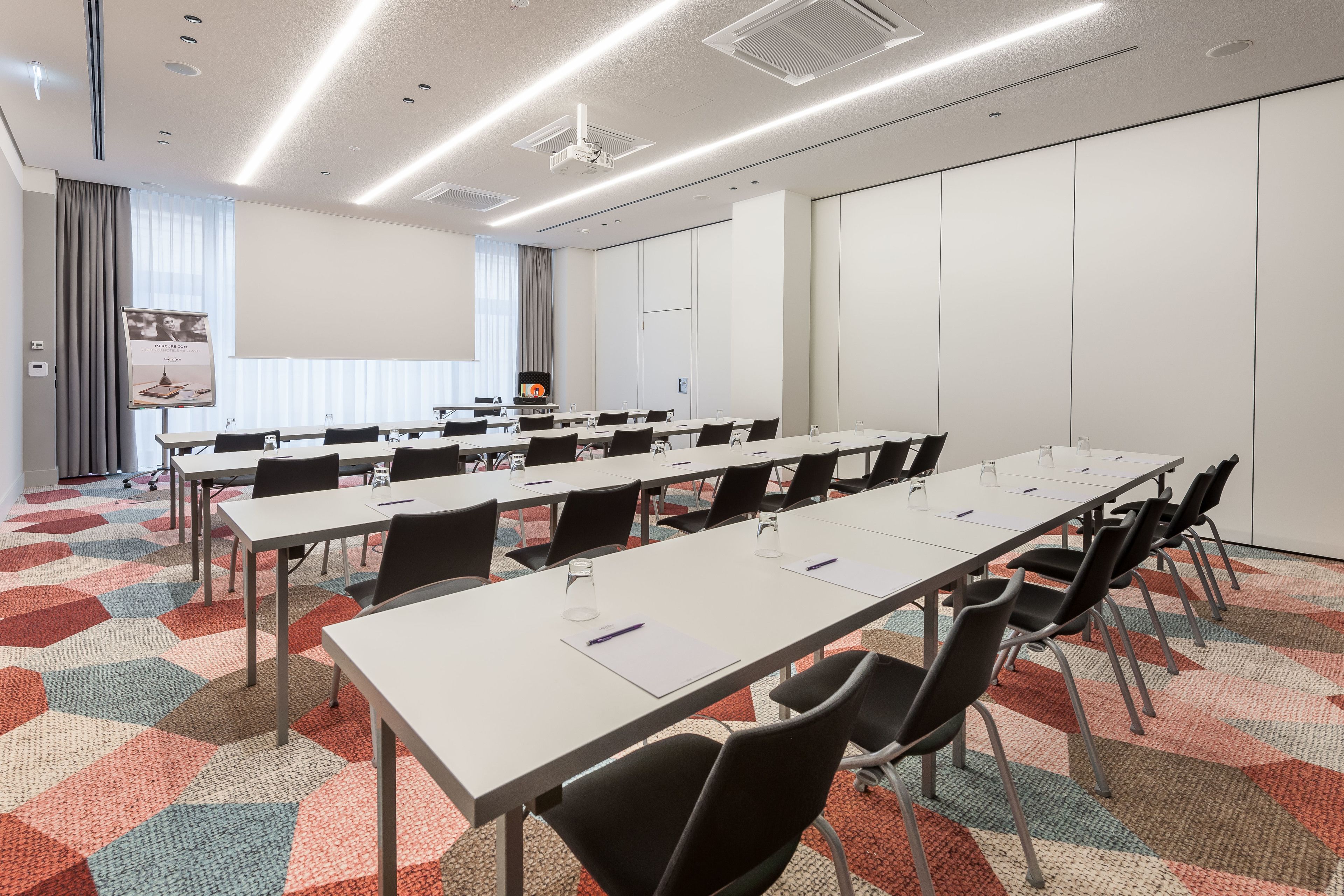 Ein Konferenzraum mit langen Tischen, schwarzen Stühlen und einem farbenfrohen, geometrischen Teppich.