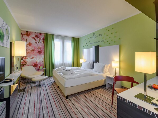 Habitación de hotel con cama, dos mesitas de noche con lámparas, un escritorio con silla, televisor y una pared con un mural floral.