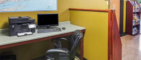 A corner office with a desktop computer, printer, and chair. There is a map on the wall and a bookshelf with magazines.