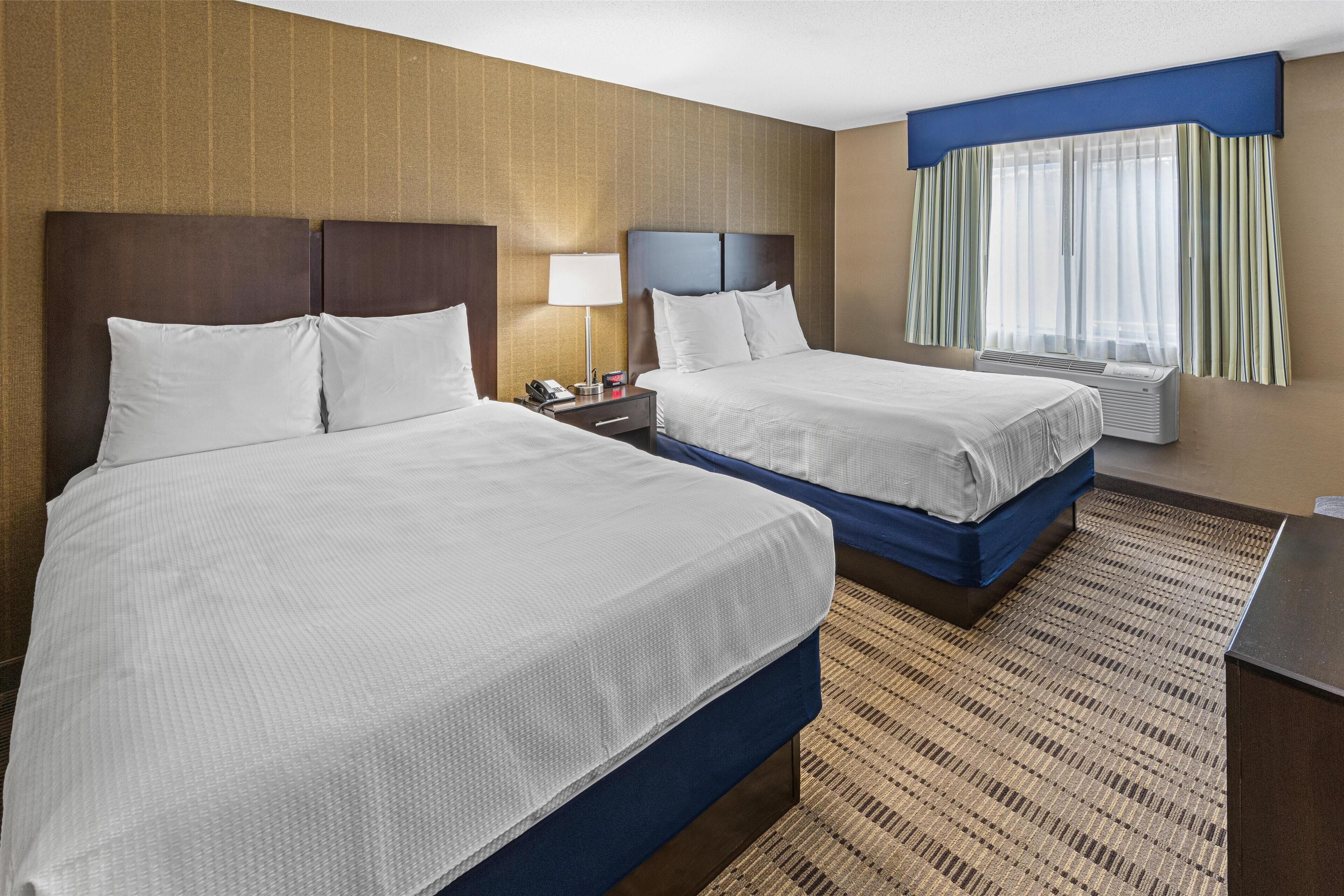 A hotel room with two beds, a nightstand with a lamp, a desk, and a window with curtains.