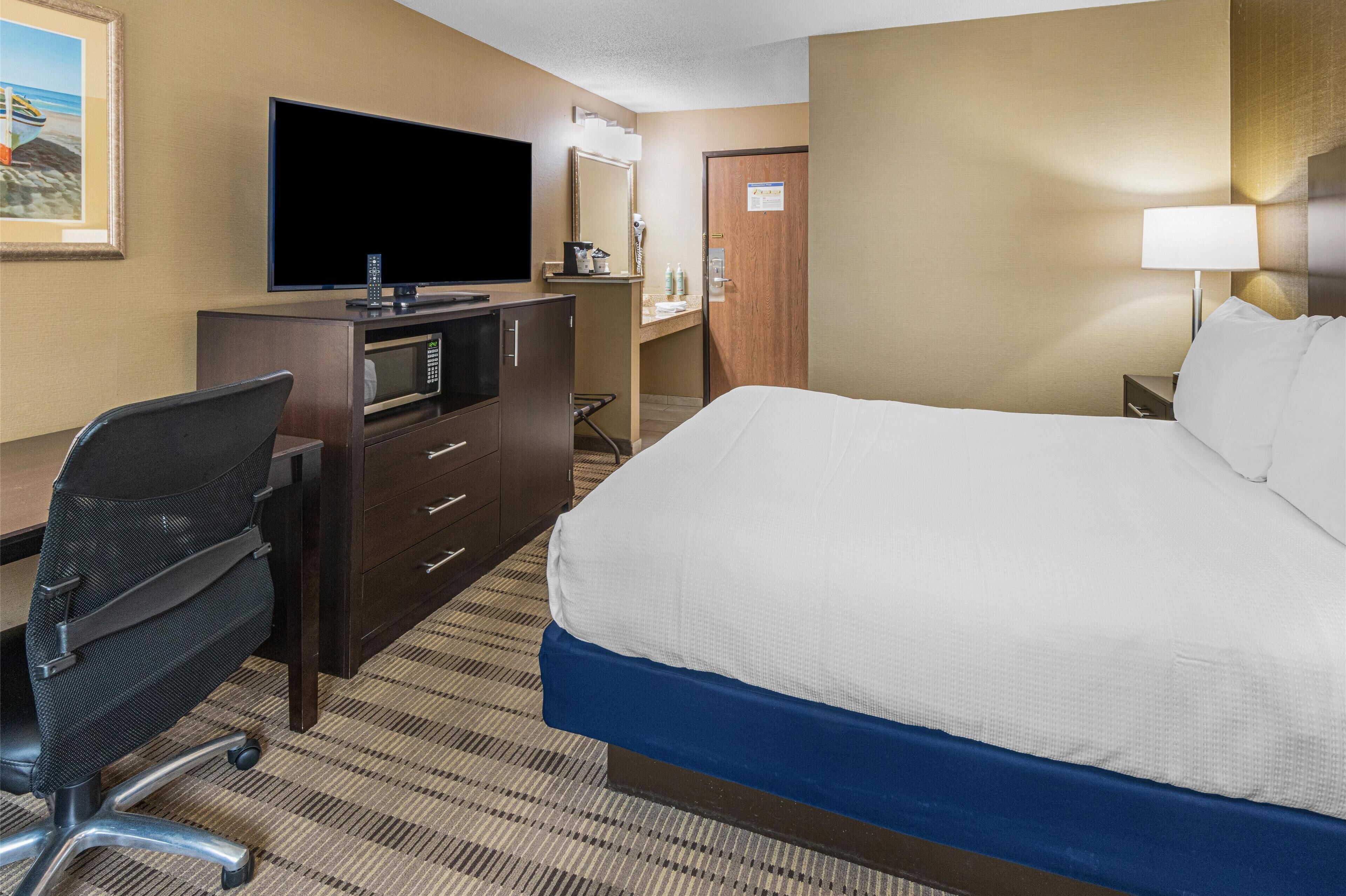 A hotel room with a bed, a desk with a flat-screen TV, a microwave, a chair, and a nightstand with a lamp.