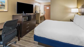 A hotel room with a bed, a desk with a flat-screen TV, a microwave, a chair, and a nightstand with a lamp.