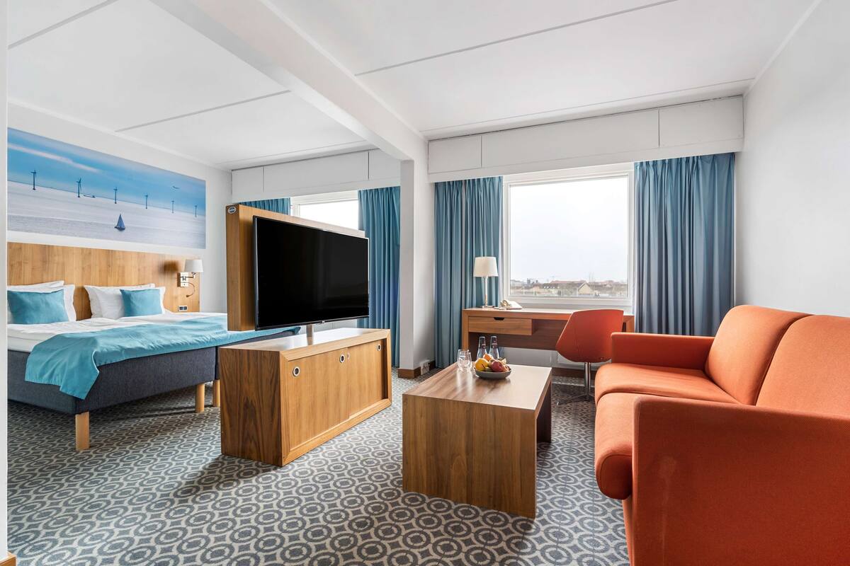 Une chambre d’hôtel équipée d’un lit, d’une télévision, d’un bureau, d’un canapé et d’une grande fenêtre avec des rideaux.