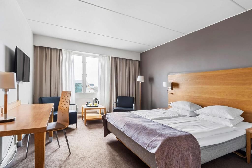 Habitación de hotel con cama, escritorio, silla, televisor y ventana con cortinas.