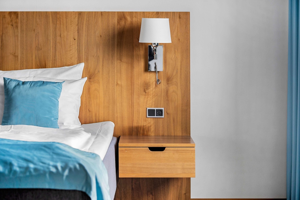Une tête de lit en bois, une lampe fixée au mur, une table de chevet avec un tiroir et un lit avec une literie bleue et blanche.
