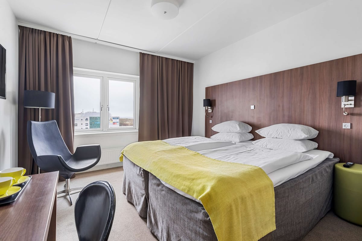 Une chambre d’hôtel avec un lit, un bureau et une chaise, une lampe, et une vue sur la ville par la fenêtre.