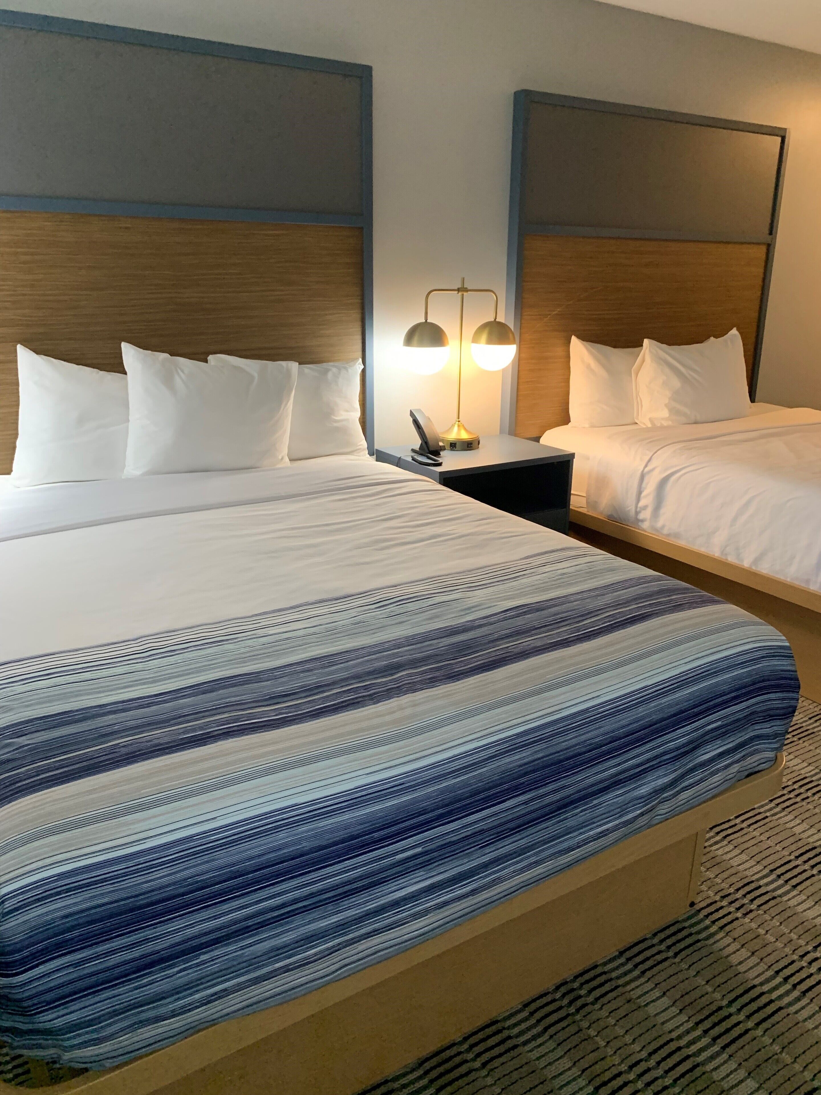 Habitación de hotel con dos camas, cada una con ropa de cama y almohadas blancas. Hay una mesita de noche con una lámpara entre las camas.