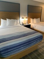 Habitación de hotel con dos camas, cada una con ropa de cama y almohadas blancas. Hay una mesita de noche con una lámpara entre las camas.