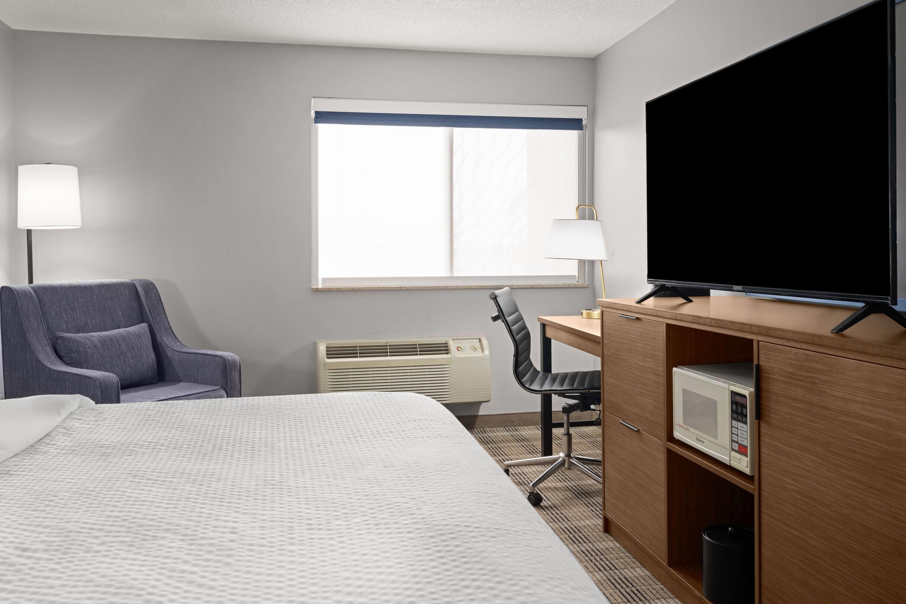 A hotel room with a bed, a desk with a television, a chair, a lamp, and a microwave.