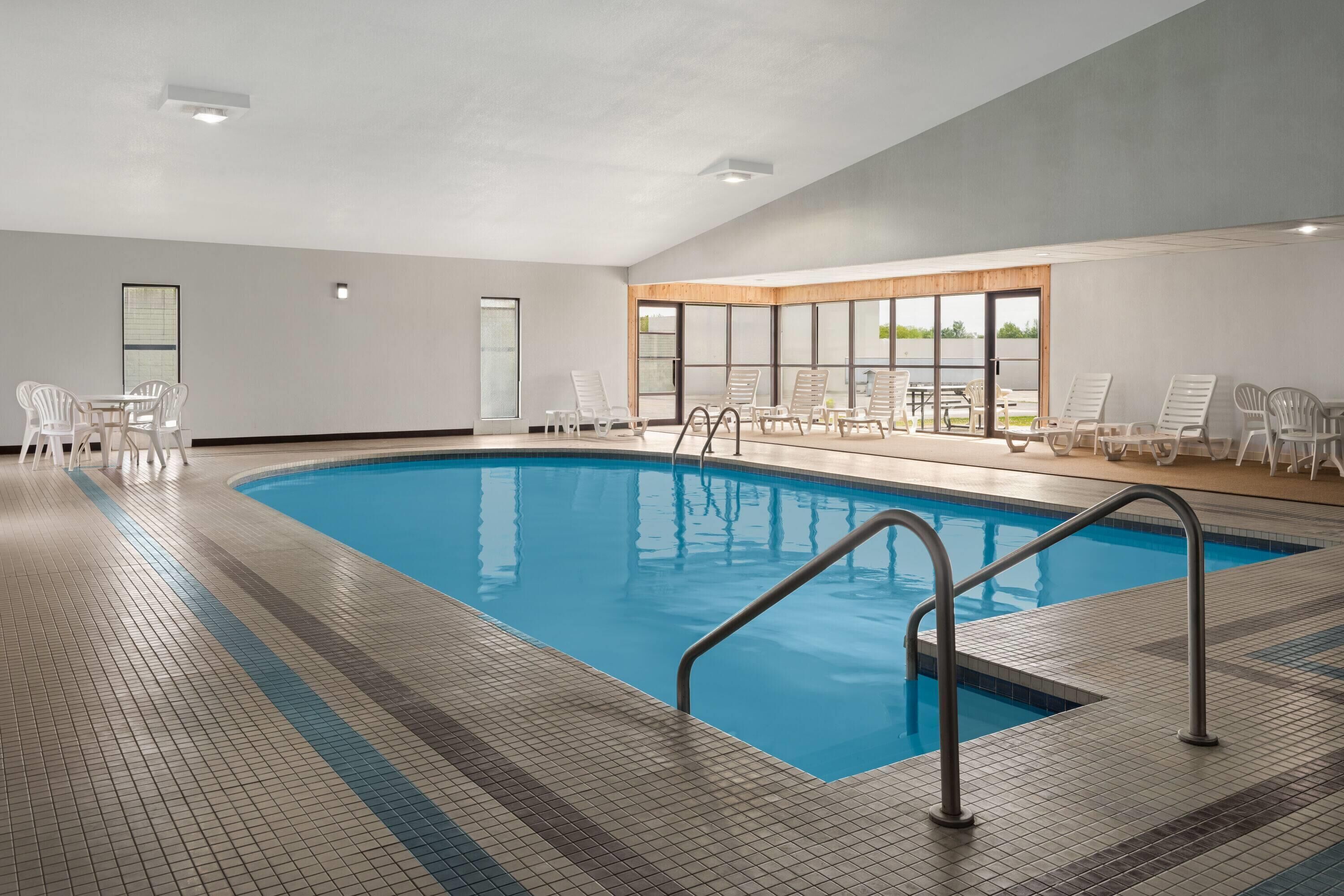 A swimming pool with a tiled deck, lounge chairs, and a view of the outdoors through large windows.