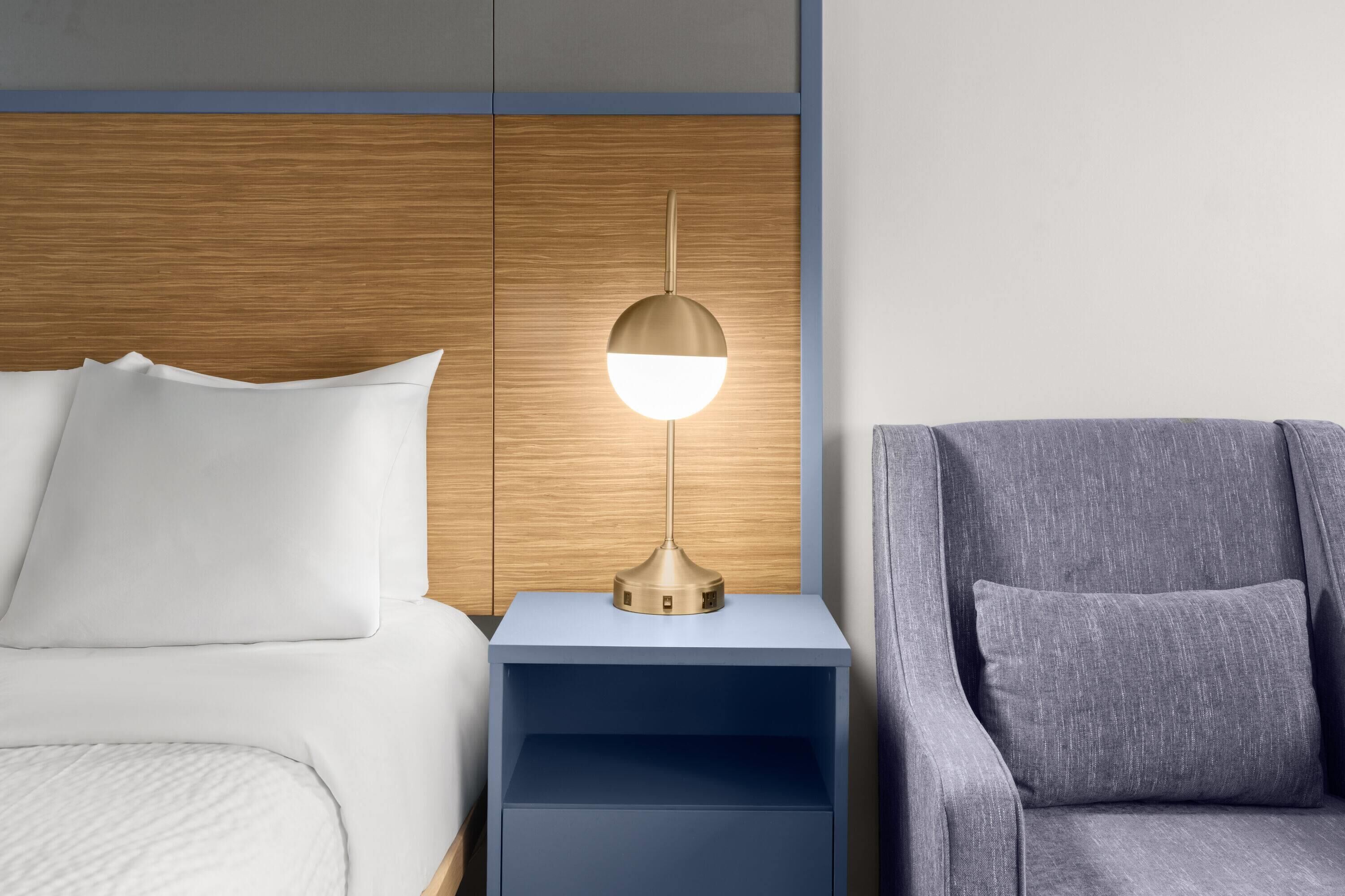 A bed with white bedding, a bedside table with a lamp, and a grey armchair.