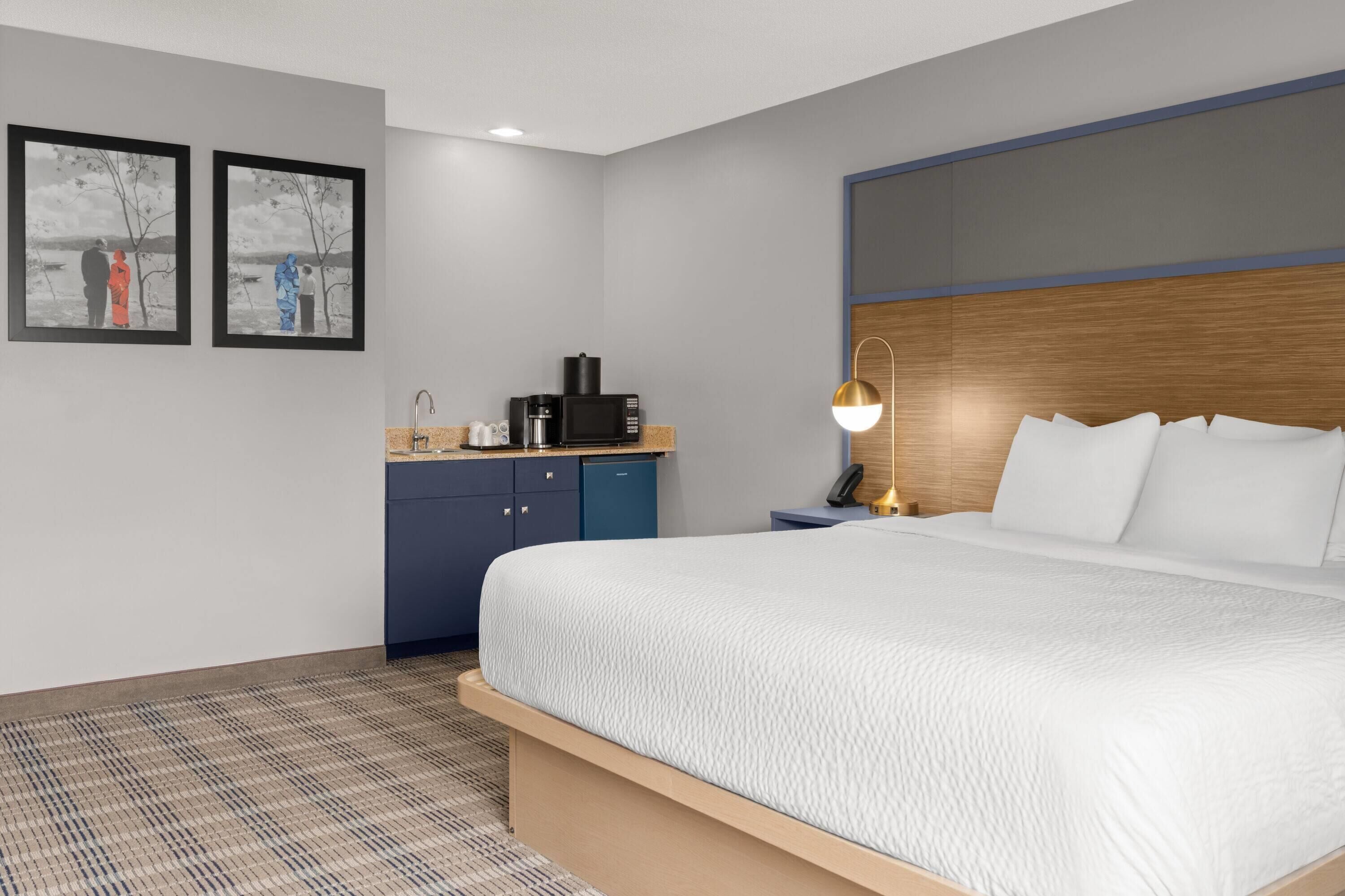 Habitación de hotel con cama, escritorio con cafetera, lámpara y dos cuadros en la pared.
