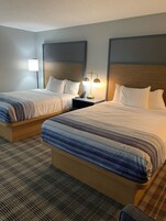 Habitación de hotel con dos camas, una mesita de noche con lámpara y una alfombra estampada.